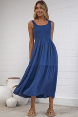 A woman modeling a sleeveless tiered midi dress in a dusty blue color, standing in a bright studio with a white brick wall background.