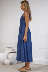 A woman stands in profile wearing a sleeveless, knee-length blue maxi dress in a bright studio with a white brick wall, light wood floor, and a beige rug. Decorative white vases sit on the floor beside her.