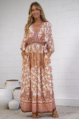 A woman stands indoors wearing a long bohemian-style maxi dress with a warm orange, rust, and cream paisley pattern. The dress features a V-neckline, elbow-length sleeves, and a gathered waist.