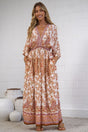 A woman stands indoors wearing a long bohemian-style maxi dress with a warm orange, rust, and cream paisley pattern. The dress features a V-neckline, elbow-length sleeves, and a gathered waist.