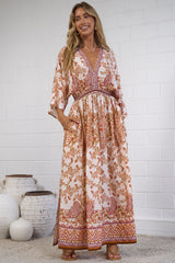 A woman modeling a bohemian-style maxi dress with a rust-orange and cream floral pattern, standing in front of a white brick wall with decorative white vases on the floor.