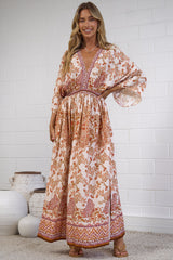 A woman modeling a bohemian-style maxi dress with a warm orange and beige floral pattern, standing in front of a white brick wall.