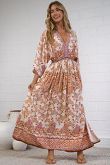 A woman modeling a bohemian-style maxi dress with a warm orange, pink, and beige floral pattern, standing in a bright room with a white brick wall and a textured rug.