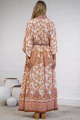 A woman stands with her back to the camera wearing a long-sleeved, ankle-length kaftan dress with an orange and brown floral pattern on a cream background. She is standing on a textured beige rug in front of a white brick wall.