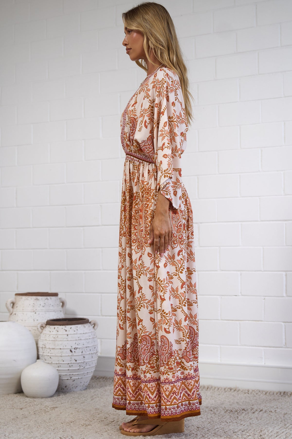 A woman stands in profile wearing a long-sleeve maxi dress with an intricate rust-orange and cream floral pattern. She is standing on a light beige woven rug against a white brick wall, with decorative white ceramic vases on the floor to her left.