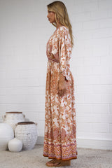 A woman stands in profile wearing a long-sleeve maxi dress with an intricate rust-orange and cream floral pattern. She is standing on a light beige woven rug against a white brick wall, with decorative white ceramic vases on the floor to her left.