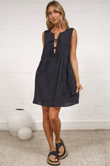 A woman modeling a sleeveless black dress with a keyhole cutout, standing on a textured beige rug against a white brick wall.