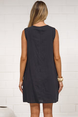 A woman stands with her back to the camera wearing a sleeveless black dress, posing in a minimalist room with a white brick wall, a beige rug, and white vases.