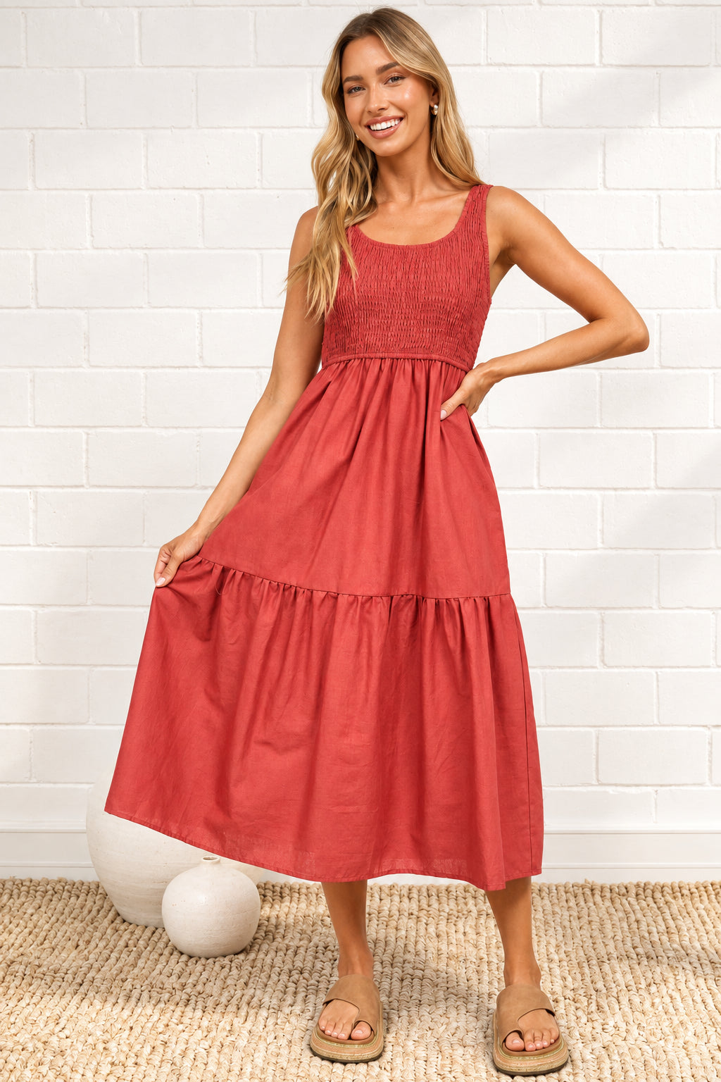 A woman modeling a sleeveless tiered midi dress in a dusty rose red color, standing on a textured beige rug against a white brick wall. 