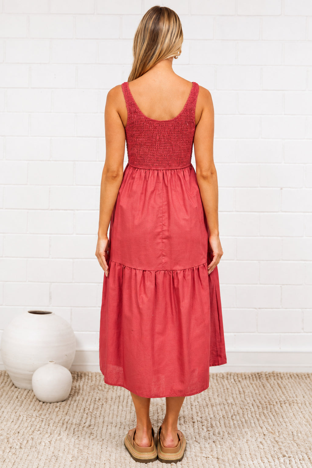 A woman stands with her back to the camera wearing a sleeveless terracotta pink tiered midi dress in a bright room with a white brick wall, a beige rug, and decorative vases. 