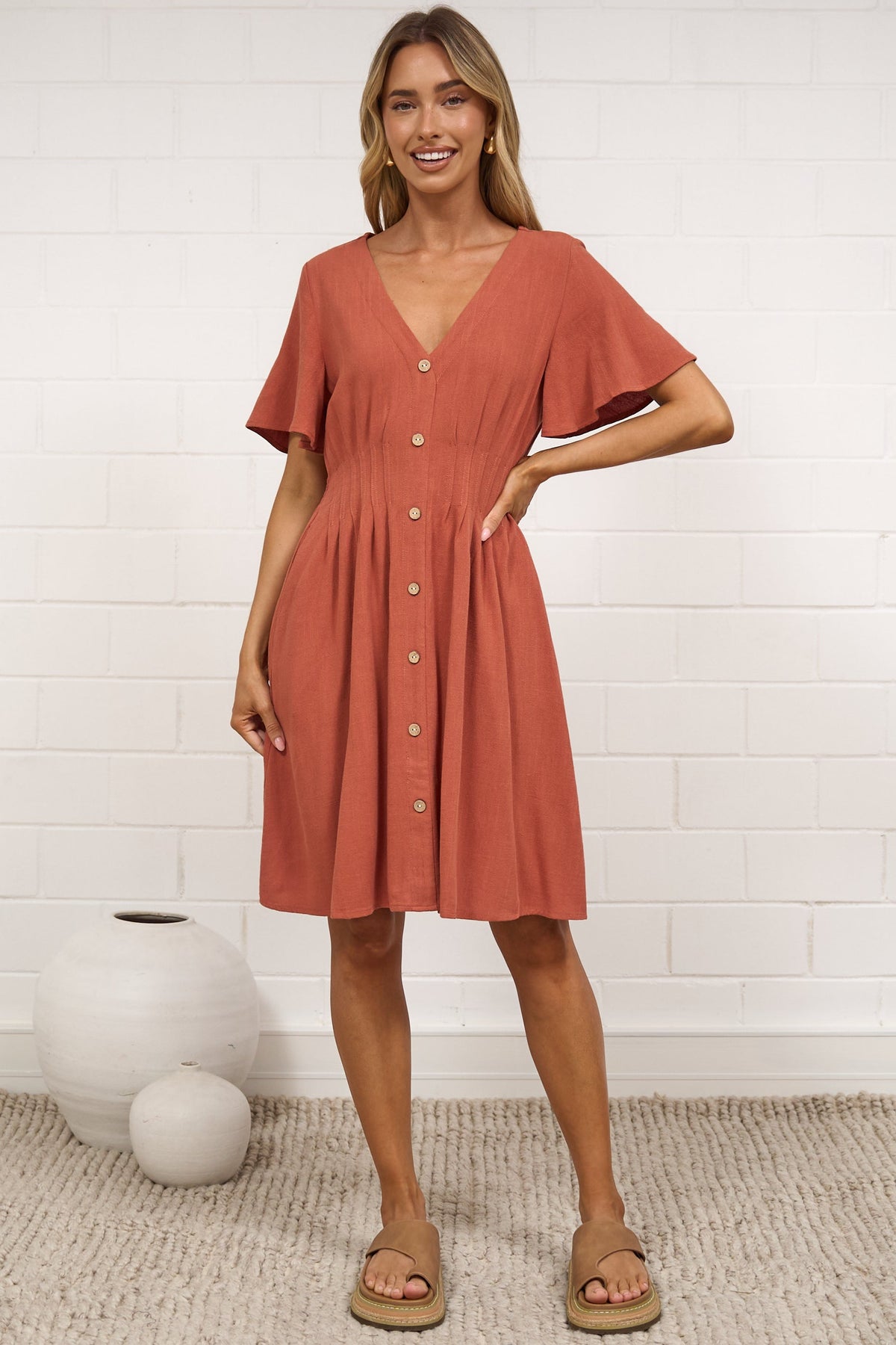 A woman stands in a bright room wearing a terracotta-colored short-sleeve button-front dress. The room has a white brick wall, a beige rug, and several white decorative vases.