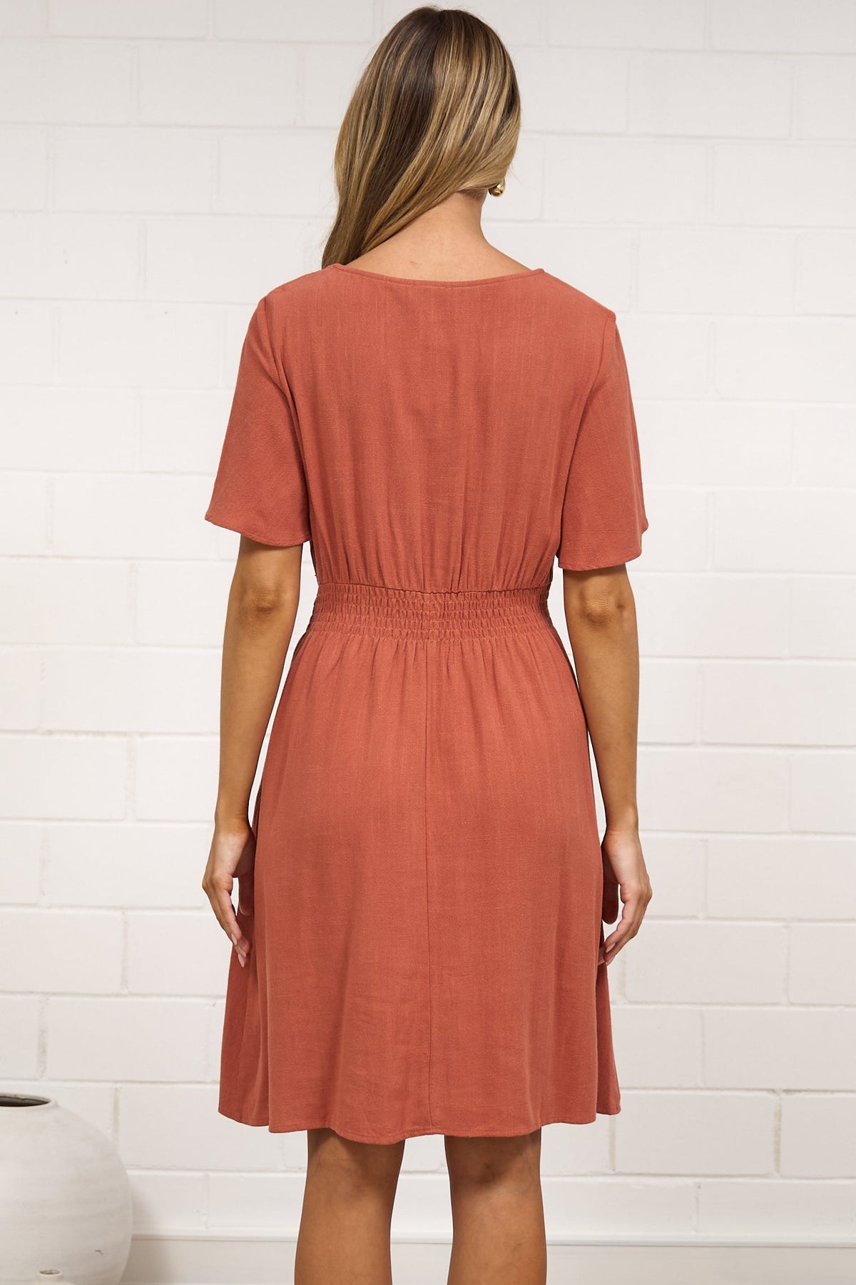A woman stands with her back to the camera wearing a short-sleeve, knee-length terracotta dress in a bright room with a white brick wall, beige rug, and white vases.