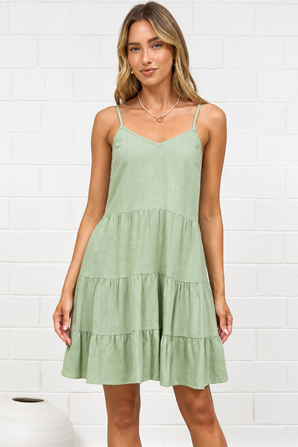 A woman modeling a light sage green tiered sleeveless dress in a bright room with a white brick wall, standing  on a beige rug.