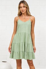 A woman modeling a light sage green tiered sleeveless dress in a bright room with a white brick wall, standing  on a beige rug.