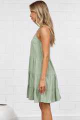 A woman stands in profile wearing a sleeveless olive green tiered dress, standing on a light beige jute rug against a white brick wall.