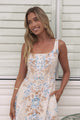 A woman wearing the Camelia midi dress in a dusty blue and tan floral print, featuring a square neckline and wide straps, standing in front of a white panelled wall.