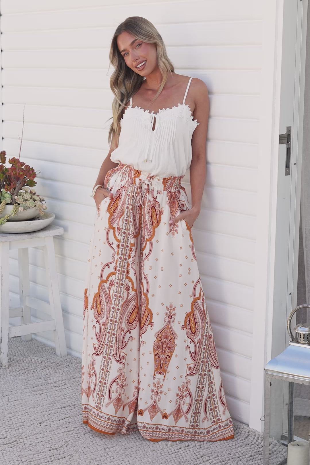 Video of a smiling woman in the Charli Pants in Sedona Print, high-waisted paper bag wide-leg pants in a cream, rust and orange paisley print, styled with a white ruffle cami against a white wall