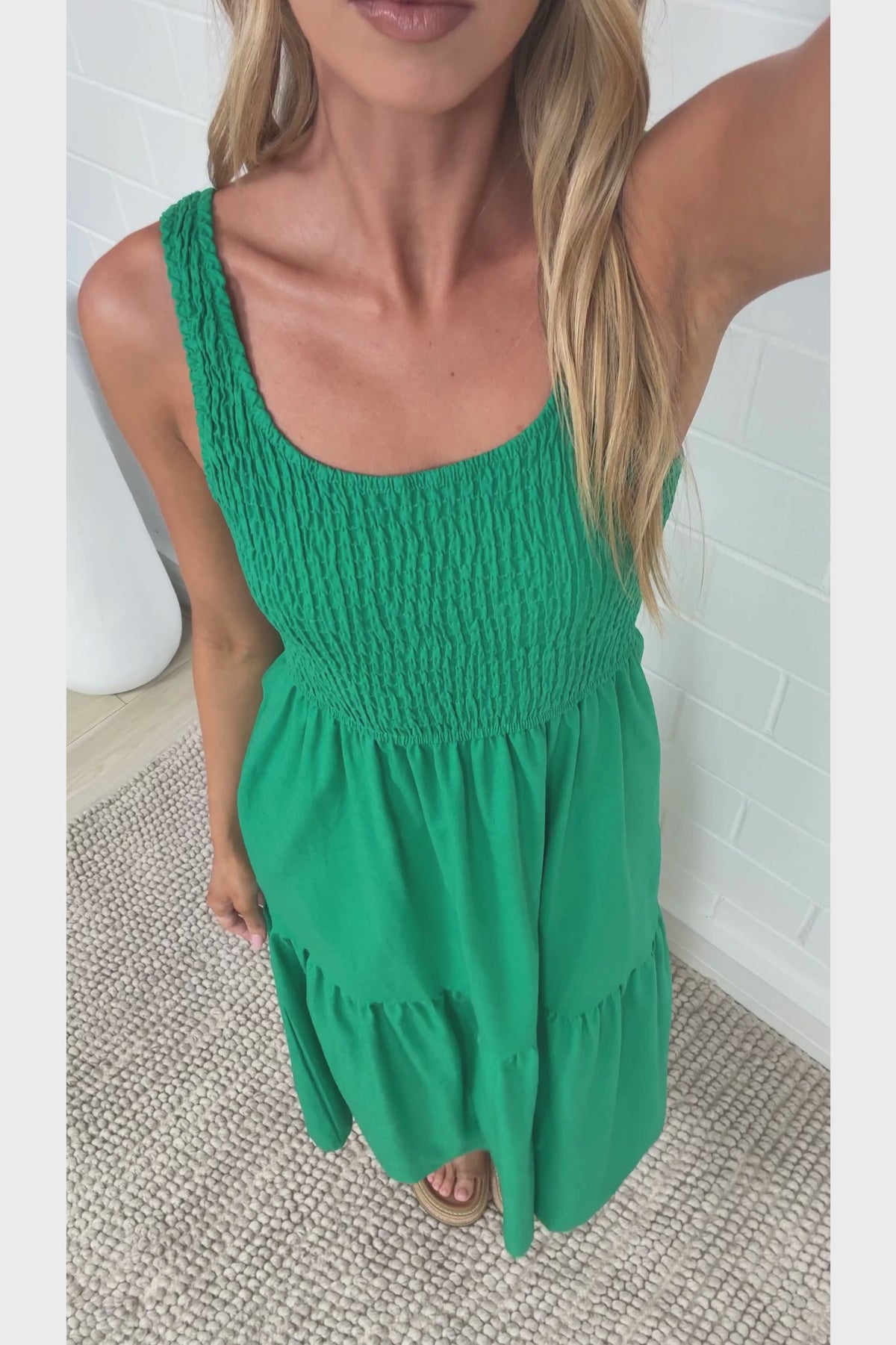 A woman in a flowing green midi dress featuring a shirred bodice and tiered skirt, posed against a white painted brick wall.