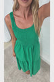 A woman in a flowing green midi dress featuring a shirred bodice and tiered skirt, posed against a white painted brick wall.