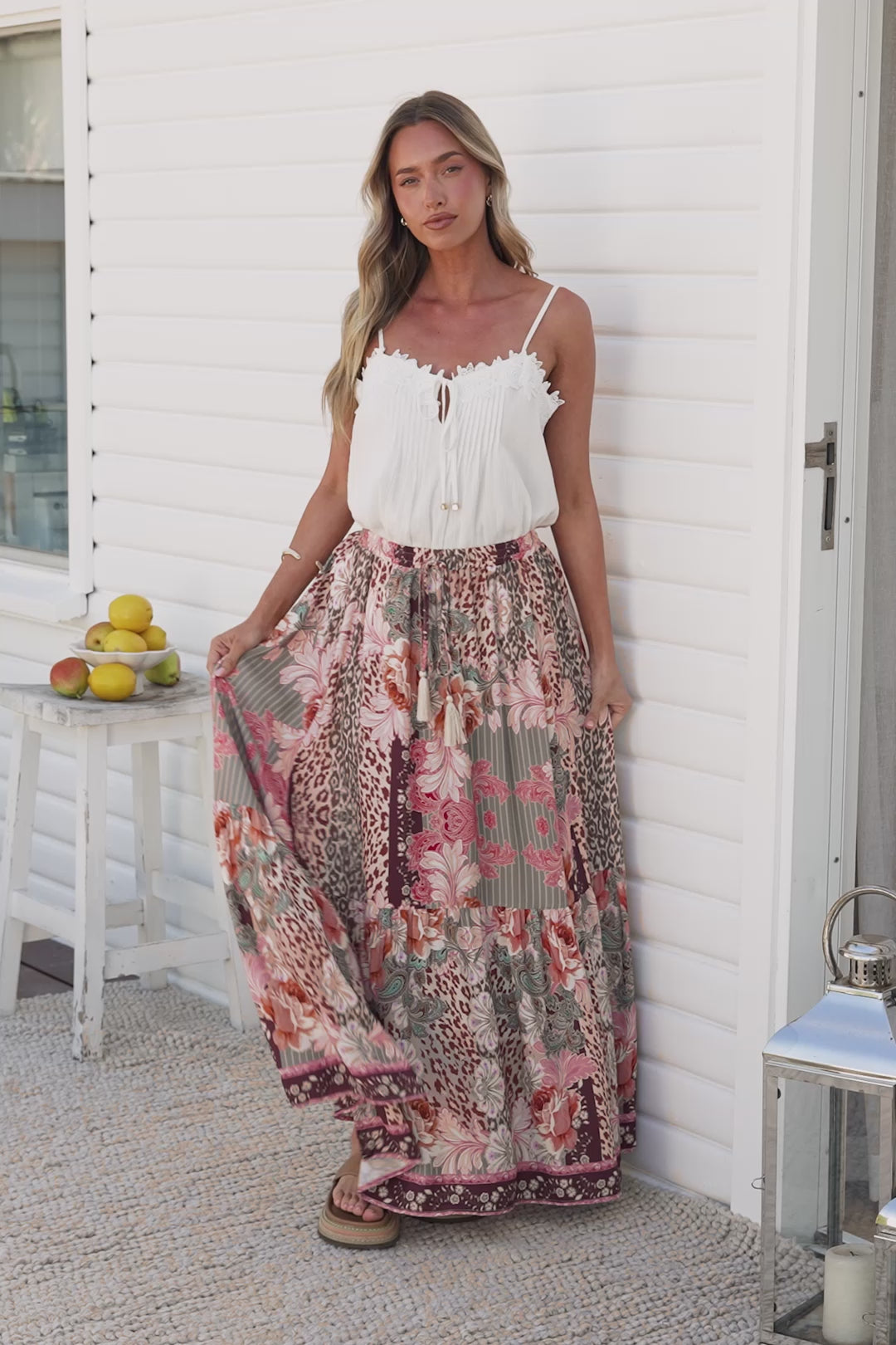 Video of a woman in the Zia Maxi Skirt in Frannie Print, a high-waisted maxi skirt in a pink, sage and leopard floral patchwork print with a tassel waist tie, styled with a white cami on a sunny porch