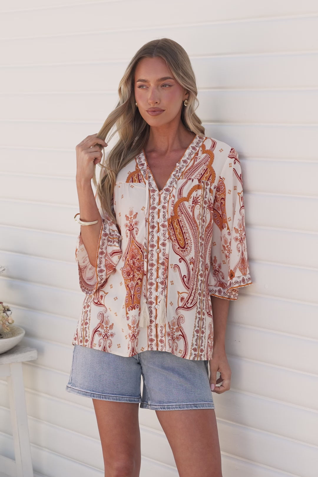 Video of a woman touching her hair in the Elysia Blouse in Sedona Print, a cream button-down with a deep V-neck, 3/4 sleeves and rust and orange paisley print, paired with light denim shorts against a white wall