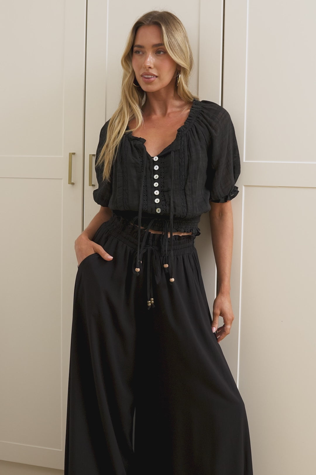Wearing high-waisted black pants with a gathered paper-bag waist and wide-leg fit, a woman stands in front of a warm beige closet.
