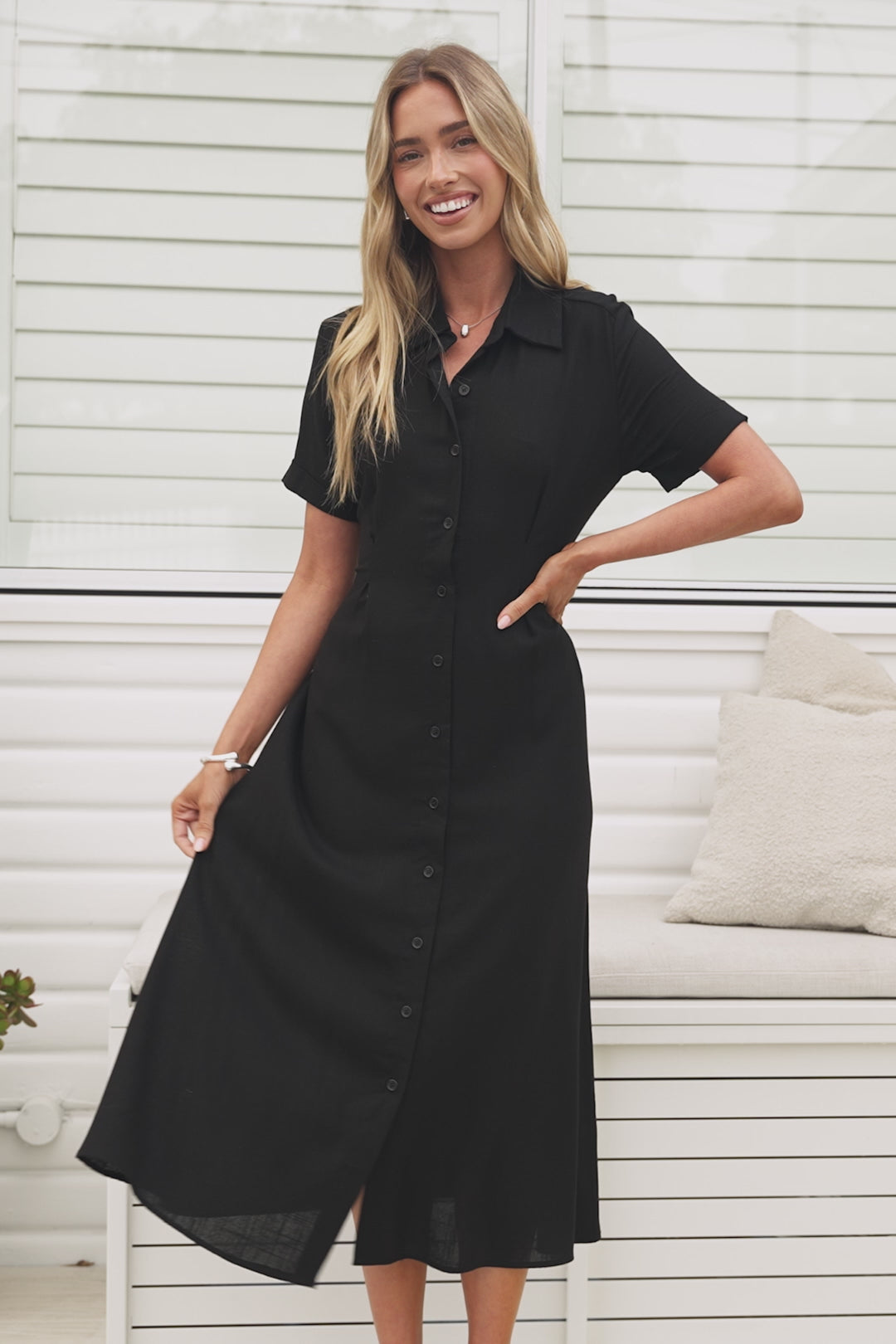 A woman wearing a black midi dress with a round neckline, short sleeves, and relaxed flowing silhouette, standing in front of a white panelled wall.